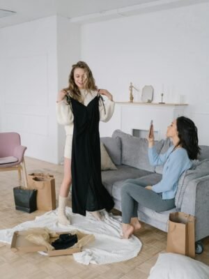 Two women trying on dresses in a stylish living room, surrounded by shopping bags and cozy decor.